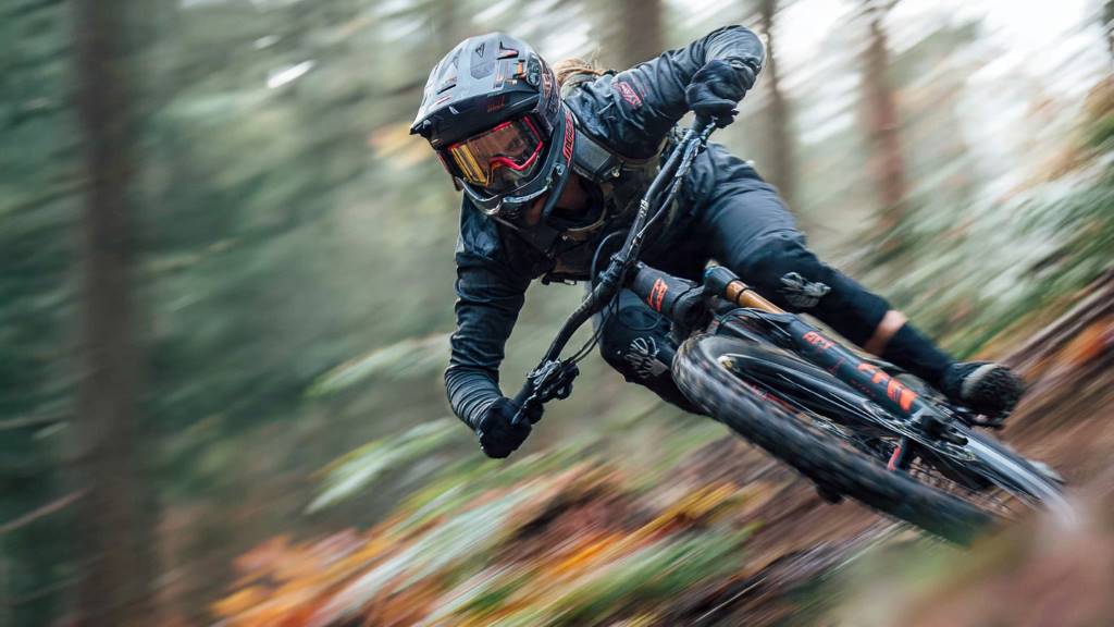 Rider wearing a full-face MTB helmet descending a steep, forested singletrack, with motion blur emphasizing speed and protection.